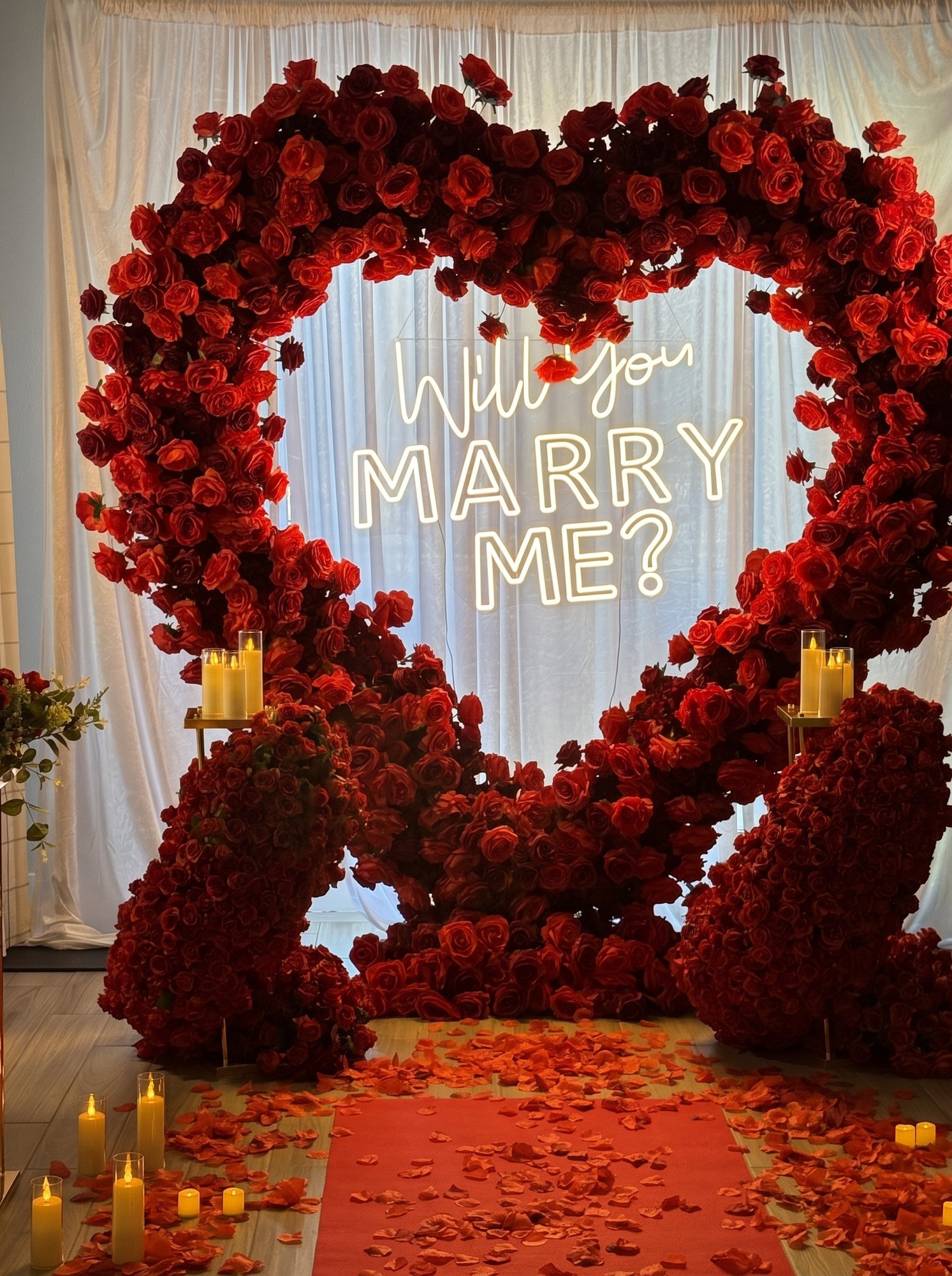 A proposal set-up: a heart-shaped arch built entirely from deep red roses framing a glowing Will You Marry Me neon sign, pillar candles either side and a carpet of scattered petals leading up to it.