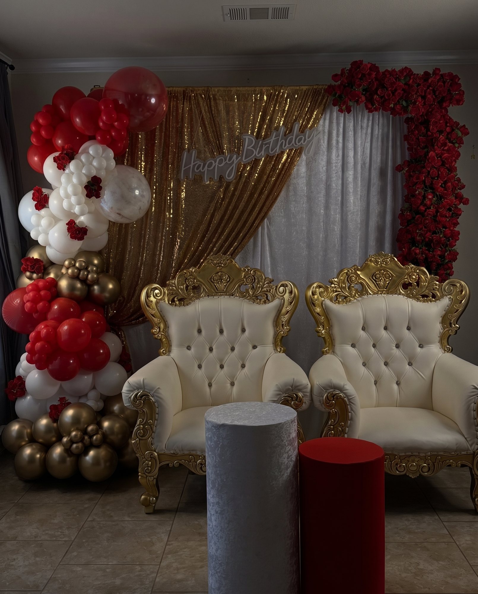 Two carved gold throne chairs under a lit backdrop, framed by a crimson, gold and ivory balloon garland with oversized florals.