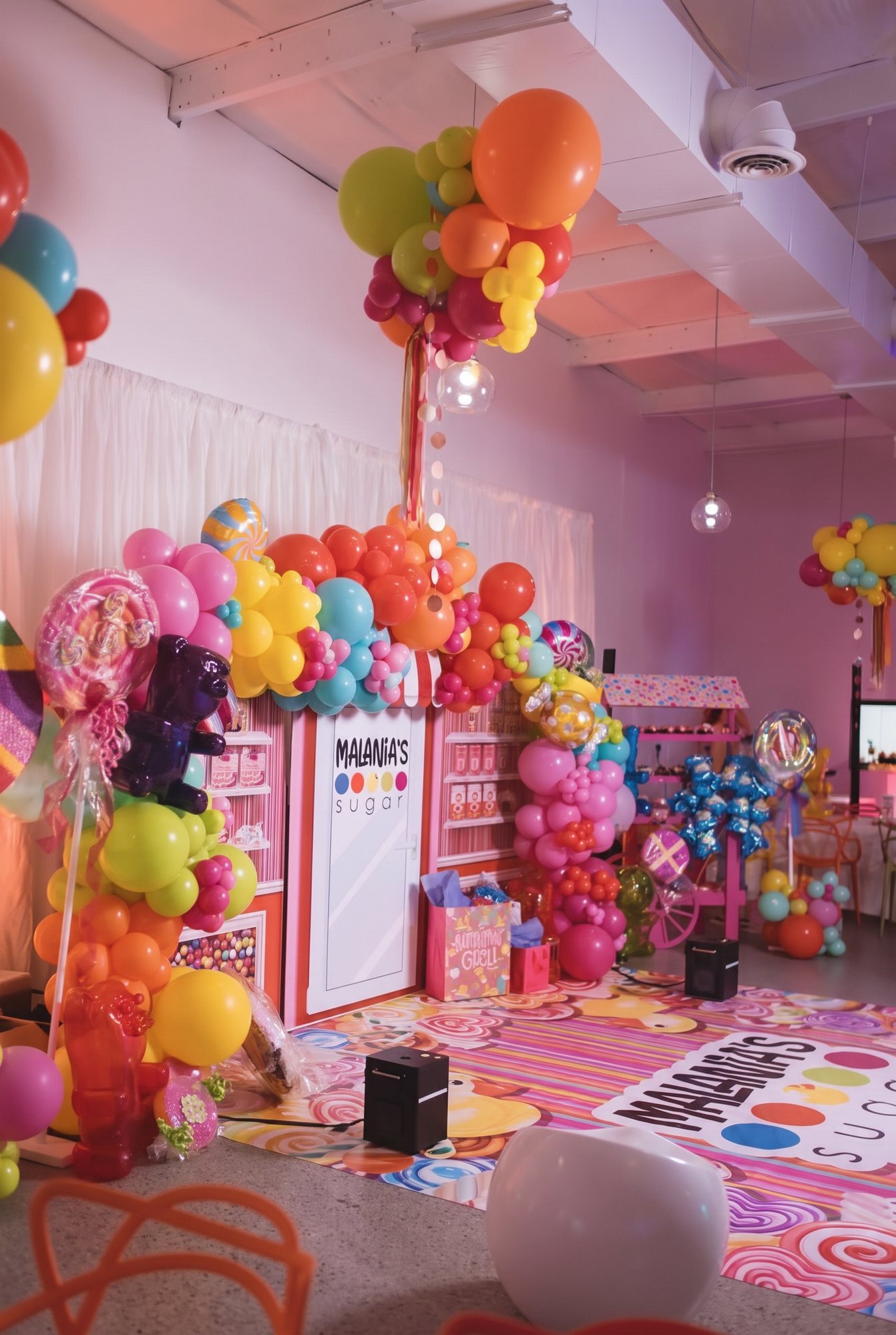 A sweet-shop themed party room styled end to end: a tall balloon column in rainbow brights, a dressed candy counter and a patterned floor wrap running the length of the space.