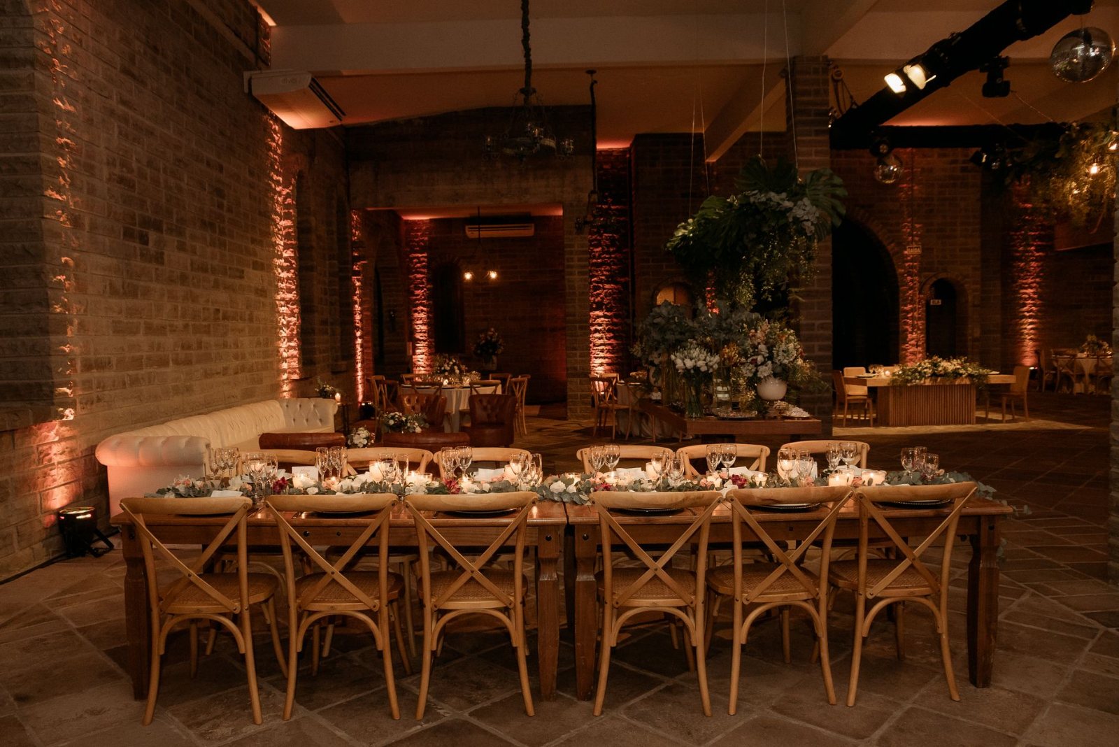 A styled table and seating setup dressed for a celebration — linen runner, upholstered chairs, charger plates, low floral centrepiece and taper candles lit, shot at a low angle down the length of the table in warm ambient light