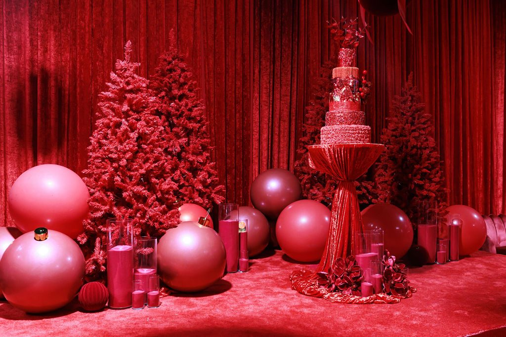 An all-red winter scene: flocked crimson trees, a tiered cake on a tall pedestal and clusters of oversized pink and red spheres on a red carpeted floor.