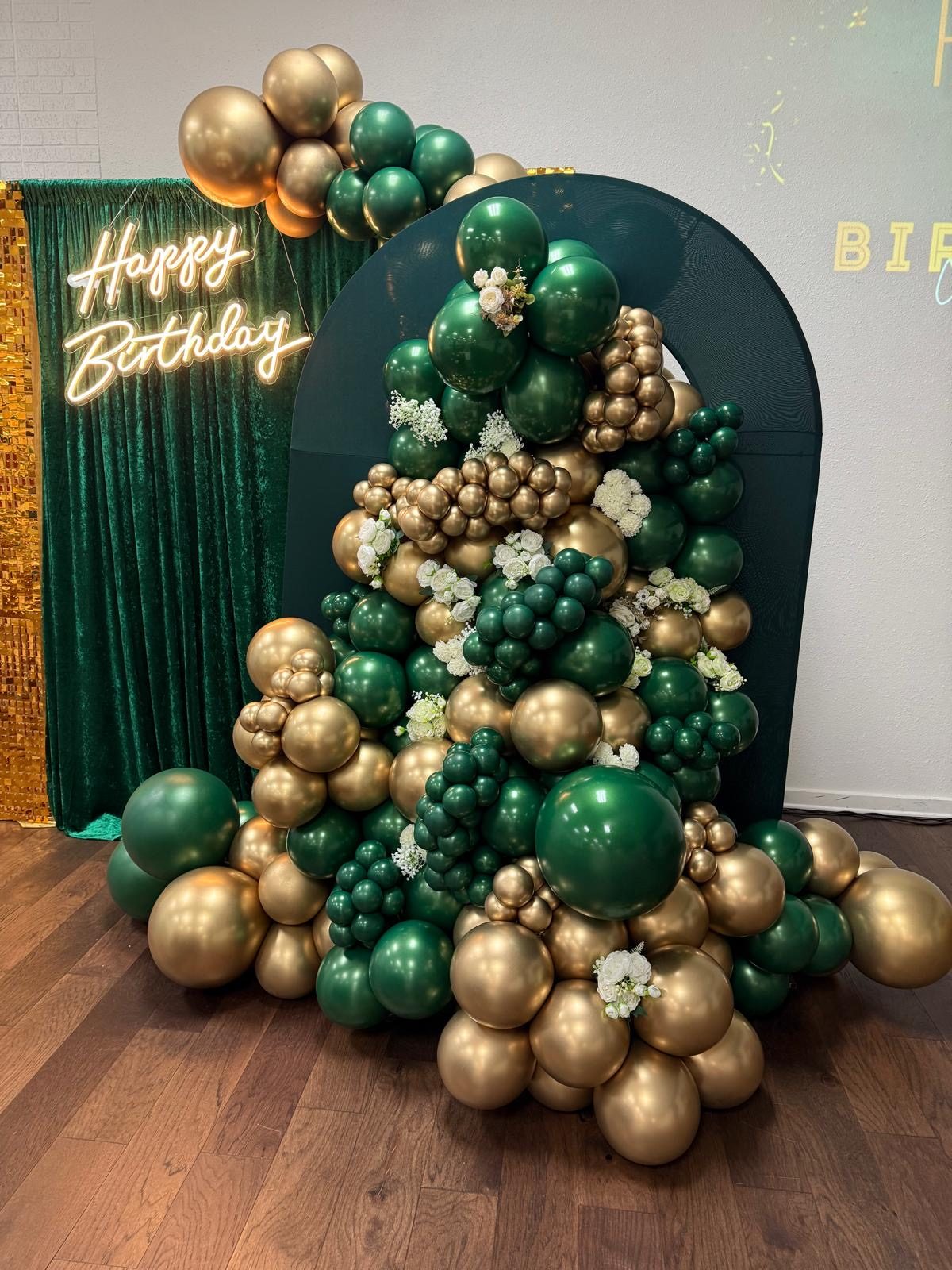 An emerald and gold birthday backdrop: a rounded arch panel with a Happy Birthday neon script, a sequin drape and a cascading balloon garland in forest green, gold chrome and ivory.