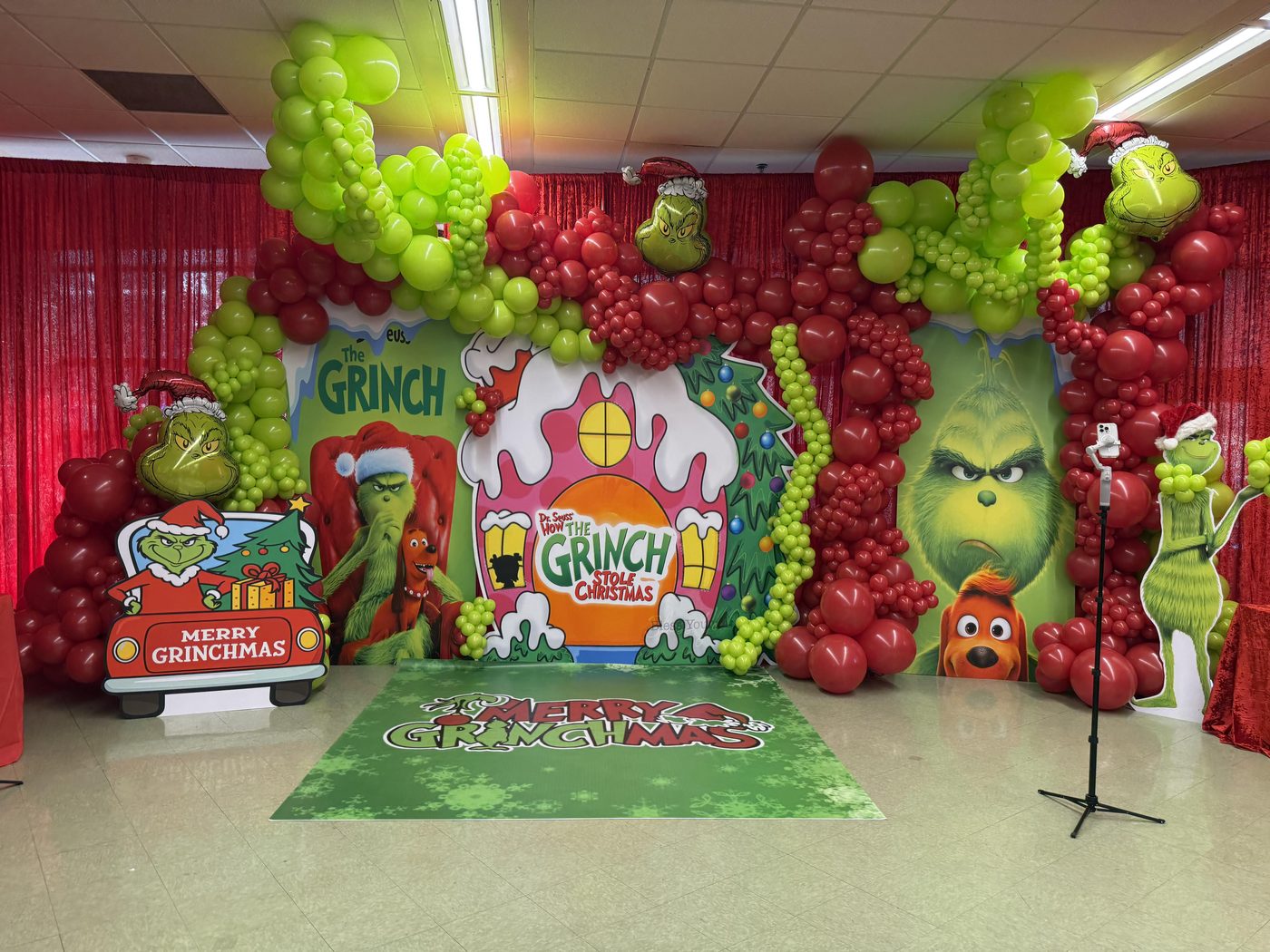 A Grinch themed Christmas set with a painted house backdrop, character cut-outs and green and red balloon columns either side of a printed floor wrap.